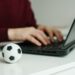 Young man reading soccer news or writing football article on laptop. Soccer ball on the table. Betting, gambling concept.